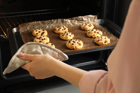 Woman taking out delicious chocolate chip cookies from oven, closeup