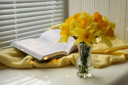 Beautiful yellow daffodils in vase, book and festive lights on windowsill. Space for textの写真素材