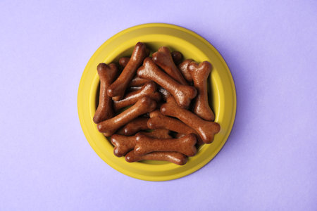 Yellow bowl with bone shaped dog cookies on purple background, top view