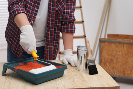 Man taking orange paint with roller from tray at wooden table indoors, closeupの写真素材
