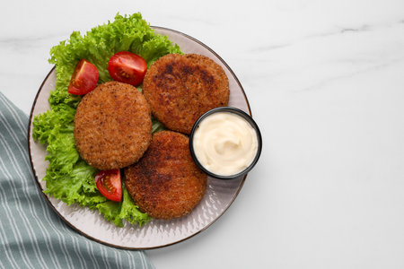 Plate with delicious vegan cutlets, lettuce, tomatoes and sauce on white marble table, top view....
