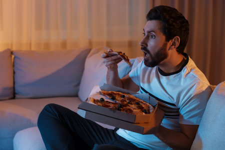 Man eating pizza while watching TV on sofa at night. Bad habitの写真素材