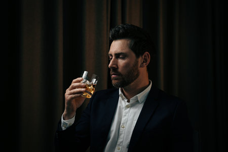 Man in suit holding glass of whiskey with ice cubes on brown backgroundの写真素材