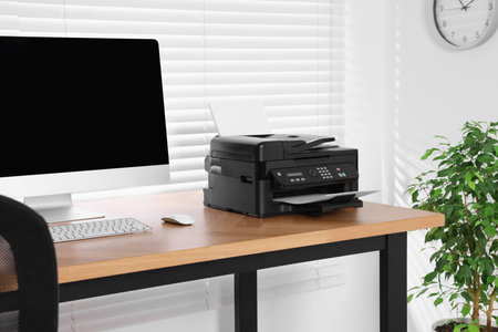 Modern printer with paper near computer on wooden table in home office
