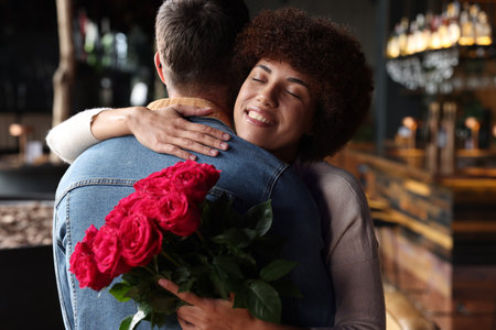 International dating. Beautiful woman with bouquet of roses hugging her boyfriend in cafeの写真素材
