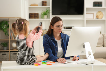 Woman working remotely at home. Mother using computer while her daughter playing with phone. Child sitting on deskの写真素材