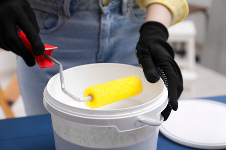 Woman dipping roller into bucket with paint at blue wooden table indoors, closeupの写真素材