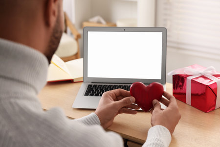 Valentine's day celebration in long distance relationship. Man holding red wooden heart while having video chat with his girlfriend via laptop, closeupの写真素材