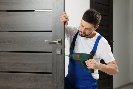 Worker in uniform with screw gun repairing door lock indoorsの写真素材