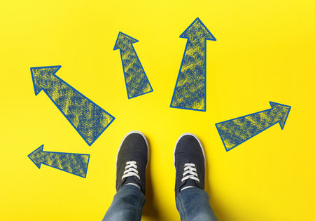 Choosing future profession. Teenager standing in front of drawn signs on yellow background, top view. Arrows pointing in different directions symbolizing diversity of opportunitiesの写真素材