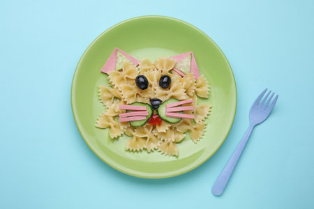 Creative serving for kids. Plate with cute cat made of tasty pasta, vegetables and sausage on light blue background, flat layの写真素材