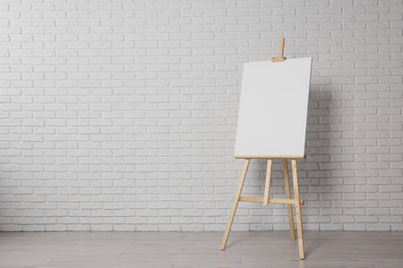 Wooden easel with blank canvas near white brick wall indoors. Space for textの写真素材