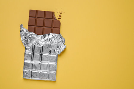 Bitten milk chocolate bar wrapped in foil on pale yellow background, top view. Space for textの写真素材