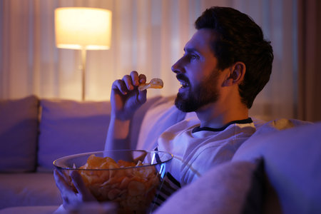 Man eating chips while watching TV on sofa at night. Bad habitの写真素材