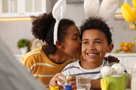 Happy African American mother kissing her cute son while painting Easter eggs at table in kitchenの写真素材