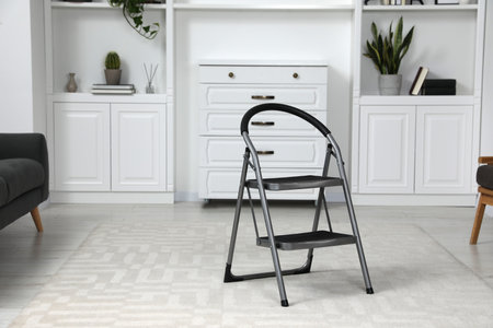 Metal folding ladder near chest of drawers and shelves with accessories in room