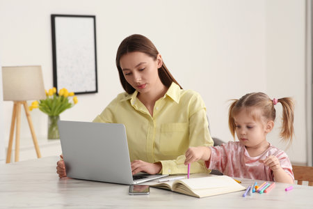 Woman working remotely at home. Mother using laptop while daughter drawing at deskの写真素材