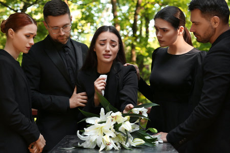Funeral ceremony. Sad people mourning near granite tombstone with white lilies at cemetery outdoors