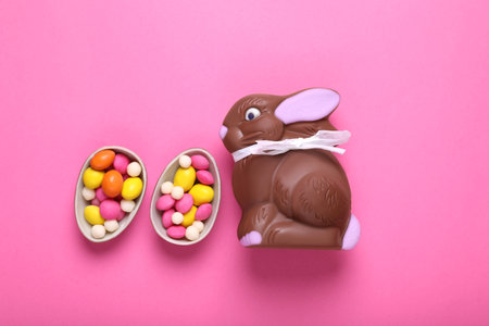 Chocolate Easter bunny, halves of eggs and candies on pink background, flat lay