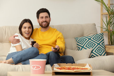 Happy couple watching TV with popcorn and pizza on sofa indoors, space for textの写真素材