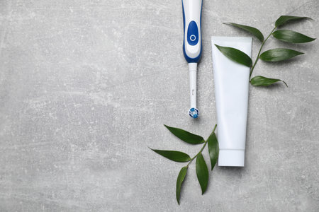 Electric toothbrush, tube of paste and leaves on gray textured table, flat lay. Space for text