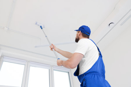 Worker in uniform painting ceiling with roller on stepladder indoors, low angle viewの写真素材