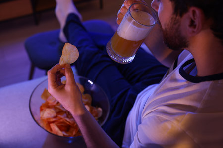 Man eating chips and drinking beer while watching TV on sofa at night, closeup. Bad habitの写真素材