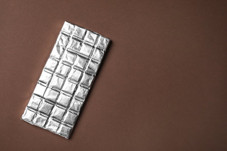 Tasty chocolate bar wrapped in foil on brown background, top view. Space for text