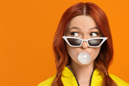 Emotional woman in sunglasses blowing bubble gum on orange background. Space for textの写真素材
