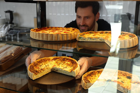 Happy seller taking tasty quiche from showcase in bakery shop