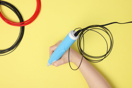 Boy drawing with stylish 3D pen on yellow background, top view
