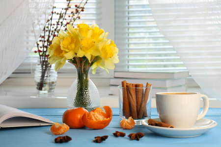 Beautiful yellow daffodils in vase, cup of coffee and tangerines on light blue wooden tableの写真素材