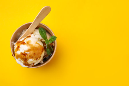 Scoops of ice cream with caramel sauce and mint leaves on yellow table, top view. Space for textの写真素材