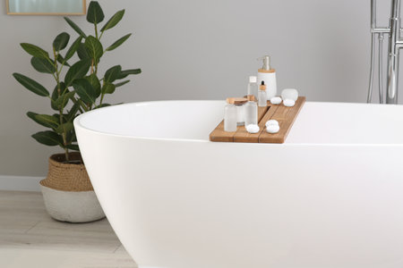 Wooden tray with cosmetic products and candles on tub in bathroom