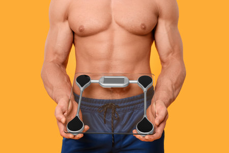 Athletic man holding scales on orange background, closeup. Weight loss concept