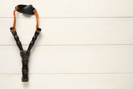 Black slingshot with pebble on white wooden table, top view. Space for text
