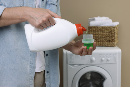Man pouring fabric softener from bottle into cap near washing machine indoors, closeupの写真素材