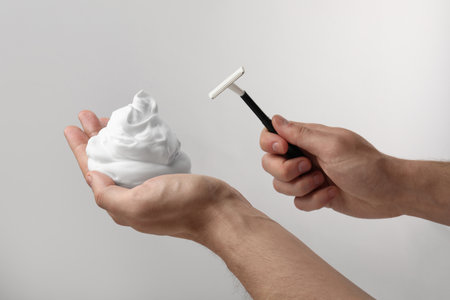Man holding shaving foam and razor on light gray background, closeup