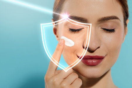 Beautiful woman applying sunscreen onto face against light blue background, space for text. Illustration of shield symbolizing sun protectionの写真素材