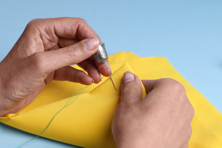 Woman sewing on yellow cloth with thimble and needle against light blue background, closeupの写真素材