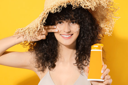 Beautiful young woman in straw hat holding tube of sunscreen against orange background
