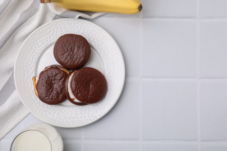 Tasty sweet choco pies, banana and glass on milk white tiled table, flat lay. Space for textの写真素材