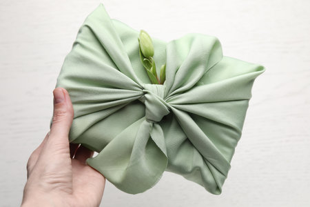 Furoshiki technique. Woman holding gift packed in green fabric with hellebore flower over light...