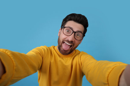 Smiling man showing tongue while taking selfie on light blue backgroundの写真素材
