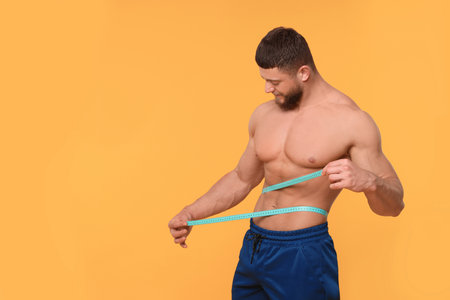 Athletic man measuring waist with tape on orange background, space for text. Weight loss conceptの写真素材