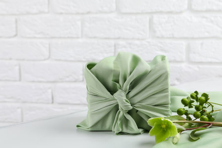 Furoshiki technique. Gift packed in green fabric and plants for decor on white table, space for textの写真素材