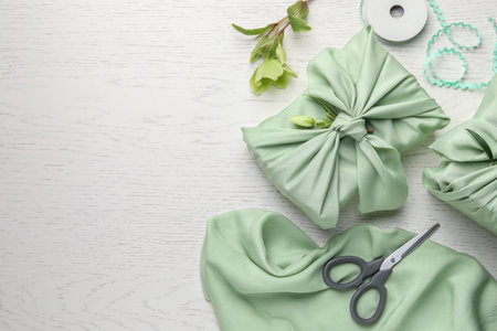 Furoshiki technique. Gifts packed in green fabrics, hellebore flowers, scissors and decorative ribbon on white wooden table, flat lay with space for textの写真素材