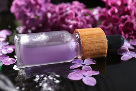 Bottle with essential oil, lilac flowers and water on black surface, closeupの写真素材