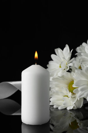 Burning candle, white chrysanthemum flowers and ribbon on black mirror surface in darkness, closeup with space for text. Funeral symbolsの写真素材