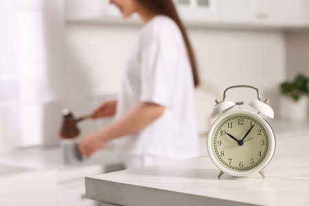 Alarm clock on white table. Woman pouring coffee from jezve into cup in kitchen, selective focusの写真素材